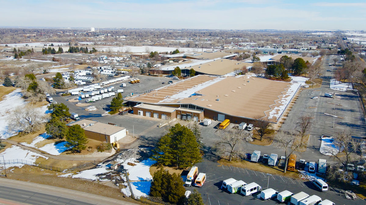 An aerial photograph of a large building partially covered in snow. There are several parking lots and a mountainscape in the background.