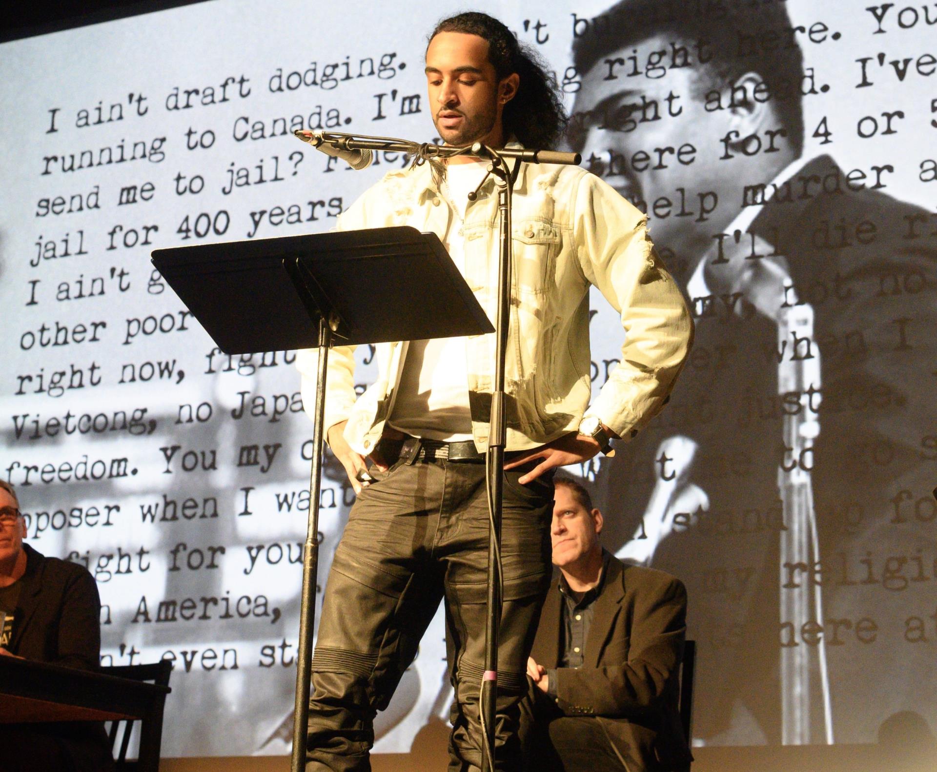 person standing on stage with a projection screen behind him and podium in front of him