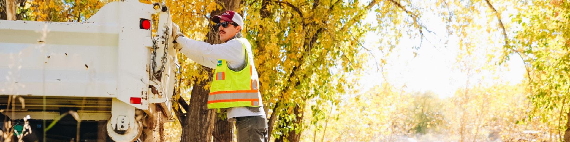 A worker leans against a truck next to a tree.