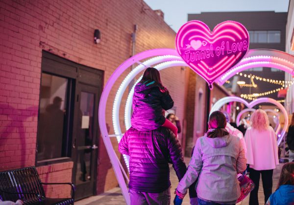 Several people walk down an alley lined with large heart arches, called the “Tunnel of Love.” A child is sitting on the shoulders of an adult.