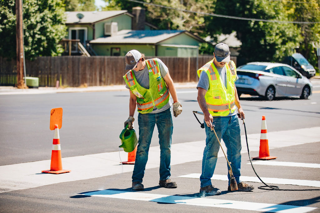 Two construction workers performing road maintenance. One worker is pouring a liquid on the road while the other is operating a tool with a long handle that is producing smoke.