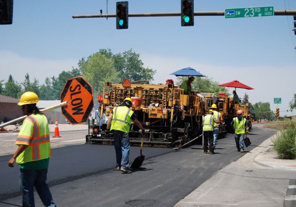 Construction workers repaving a road, with one worker holding a sign and others operating machinery under umbrellas.