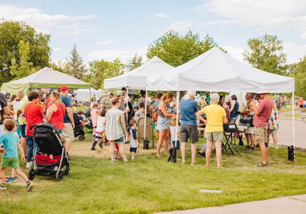 A lively outdoor event with people gathered around white tents on a sunny day, enjoying various activities.