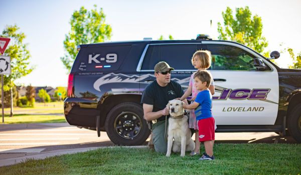 A police officer with a K-9 unit vehicle kneels with a police dog while two children pet the dog.