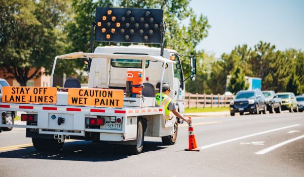 A street maintenance truck with warning signs “Stay Off Lane Lines” and “Caution Wet Paint.” A worker in a safety vest is placing an orange cone on the road.